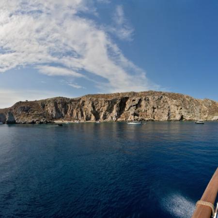 Scogliera rocciosa con barche a vela in mare blu sotto un cielo parzialmente nuvoloso.