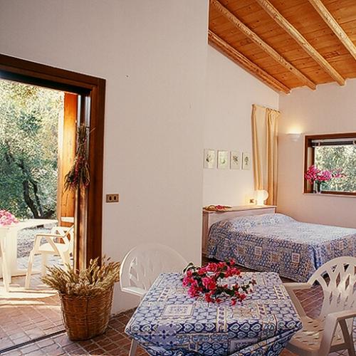 Camera rustica con letto matrimoniale, tavolo e fiori, vista su giardino esterno.